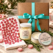 A holiday gift set from Majamas Earth featuring a kraft gift box with a turquoise ribbon, a jar of Salt Scrub, a jar of Hand & Body Cream, and a red-and-white patterned towel, arranged on a tabletop with pine sprigs and peppermint candies. 
