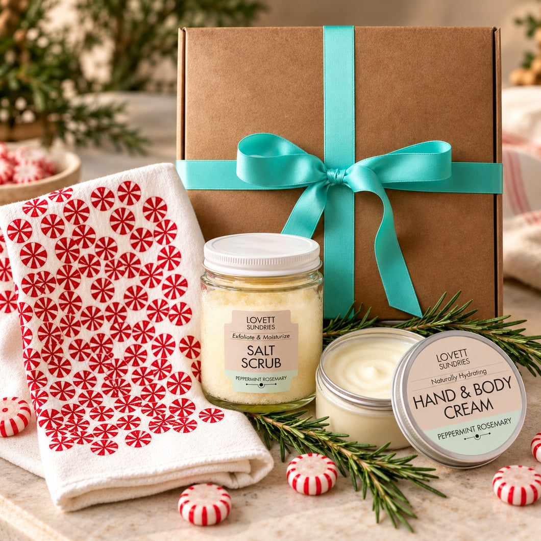 A holiday gift set from Majamas Earth featuring a kraft gift box with a turquoise ribbon, a jar of Salt Scrub, a jar of Hand & Body Cream, and a red-and-white patterned towel, arranged on a tabletop with pine sprigs and peppermint candies.