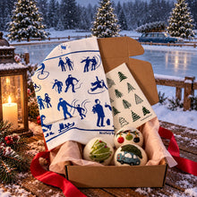 A gift box filled with winter-themed items, including a white tea towel with blue silhouettes of people sledding, a small printed card, and three decorative ornaments, set against a snowy outdoor lakeside background at dusk. 