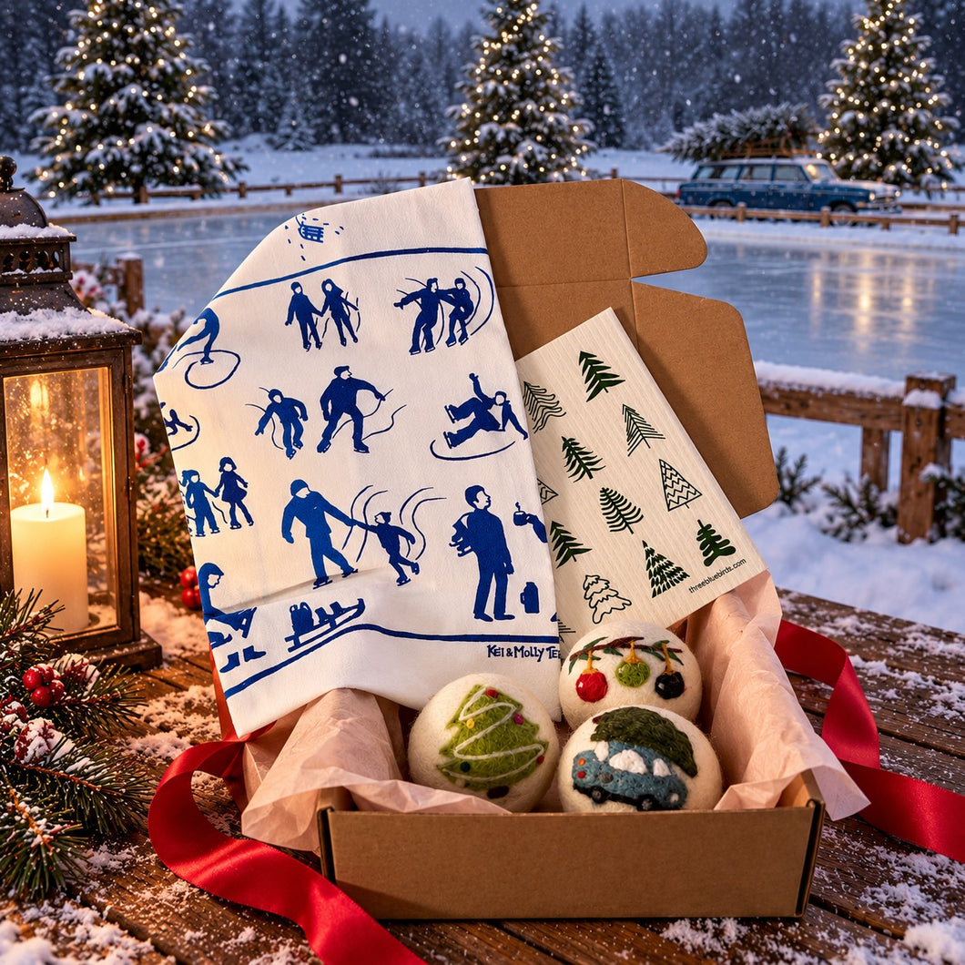 A gift box filled with winter-themed items, including a white tea towel with blue silhouettes of people sledding, a small printed card, and three decorative ornaments, set against a snowy outdoor lakeside background at dusk.