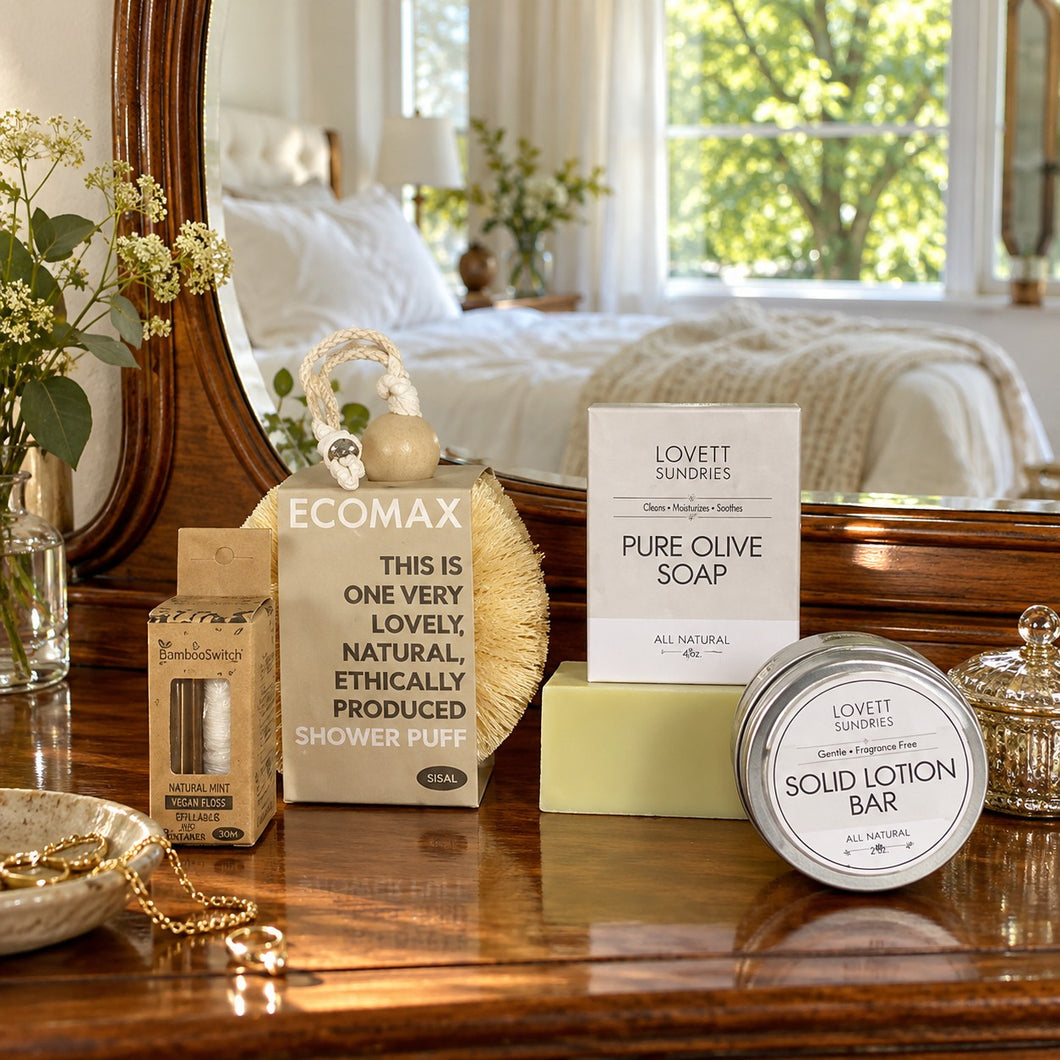 A styled bderoom scene with a wooden vanity, mirror, and soft-focus bedroom background. Visible products include a Natural Mint Bamboo Dental Floss, a Lovett Sundries Pure Olive Soap bar, a Lovett Sundries Solid Lotion Bar tin, and an Ecomax shower puff box on the vanity.